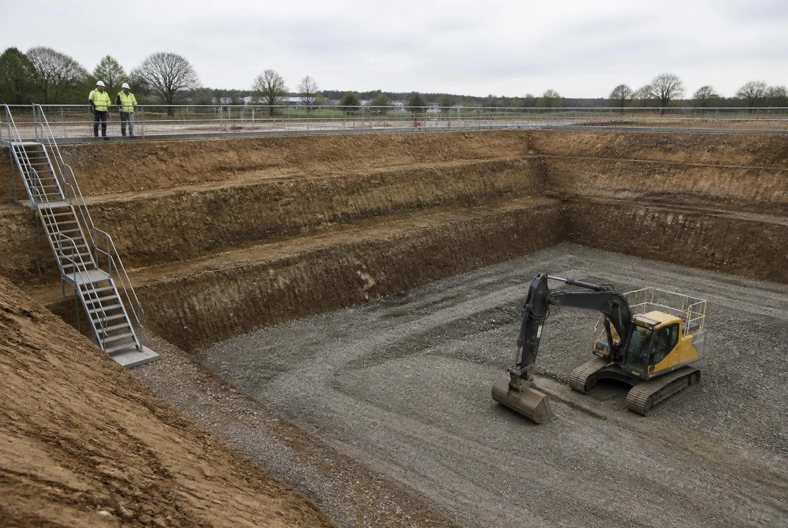 Gestufte Baugrube mit terrassierten Böschungen, Geländer und Absperrung an der gesicherten Oberkante, Treppenturm als sicherer Zugang und Kettenbagger auf der Kiesarbeitsebene am Grubenboden
